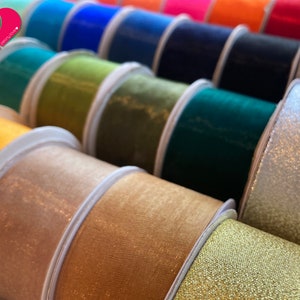 May include: A close-up of a variety of spools of ribbon in different colors and textures, including gold, silver, green, blue, and orange. The ribbons are arranged in a row, with the gold and silver ribbons in the foreground.