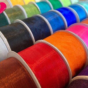 May include: A close-up of spools of colorful sheer ribbon. The ribbon is arranged in a row, with each spool a different color. The colors include red, orange, yellow, green, blue, purple, pink, and white.