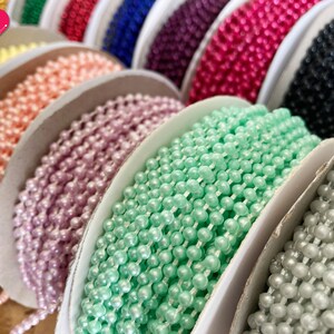 K&ouml;nnte beinhalten: Spulen mit perlenartigem Besatz in verschiedenen Farben, darunter rosa, lavendel, gr&uuml;n und wei&szlig;. Der Besatz ist um wei&szlig;e Spulen gewickelt.