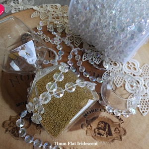 K&ouml;nnte beinhalten: Nahaufnahme eines gl&auml;sernen Sanduhr, gef&uuml;llt mit goldenem Glitzer und umgeben von regenbogenfarbenen Kristallperlen. Die Perlen befinden sich auch in einem durchsichtigen Plastikbeutel und einer Glasschale. Der Hintergrund ist eine Holzoberfl&auml;che mit einer Spitzenunterlage.