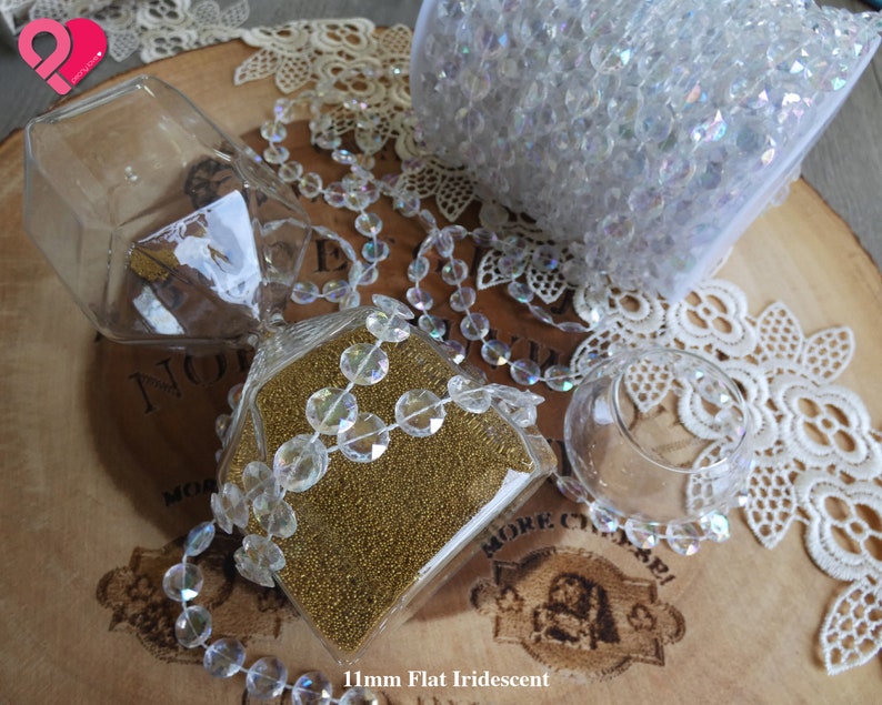 K&ouml;nnte beinhalten: Nahaufnahme eines gl&auml;sernen Sanduhr, gef&uuml;llt mit goldenem Glitzer und umgeben von regenbogenfarbenen Kristallperlen. Die Perlen befinden sich auch in einem durchsichtigen Plastikbeutel und einer Glasschale. Der Hintergrund ist eine Holzoberfl&auml;che mit einer Spitzenunterlage.