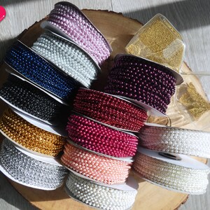 K&ouml;nnte beinhalten: Sortiment an Perlen- und Metallic-Perlenbesatz in verschiedenen Farben, darunter Silber, Gold, Wei&szlig;, Rosa, Rot, Burgunder, Blau und Schwarz. Der Besatz ist um Spulen gewickelt und auf einer Holzoberfl&auml;che ausgestellt.