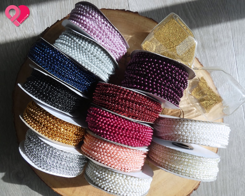 K&ouml;nnte beinhalten: Sortiment an Perlen- und Metallic-Perlenbesatz in verschiedenen Farben, darunter Silber, Gold, Wei&szlig;, Rosa, Rot, Burgunder, Blau und Schwarz. Der Besatz ist um Spulen gewickelt und auf einer Holzoberfl&auml;che ausgestellt.