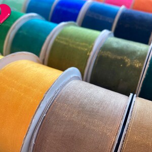 May include: A collection of spools of shimmering ribbon in various colors, including yellow, green, brown, and blue. The ribbon is made of a sheer, delicate fabric with a subtle sparkle.