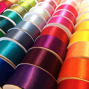 May include: Rows of colorful satin ribbon spools in various shades, including teal, pink, red, and yellow. The ribbon has a glossy finish and is neatly wound on white spools. The image showcases a variety of colors, perfect for crafting and decorative projects.