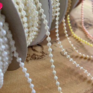 K&ouml;nnte beinhalten: Nahaufnahme von Spulen mit irisierendem Perlenbesatz in verschiedenen Farben, darunter Wei&szlig;, Rosa, Gelb und Gr&uuml;n. Der Besatz besteht aus kleinen, runden Perlen, die auf einem d&uuml;nnen Faden aufgereiht sind.