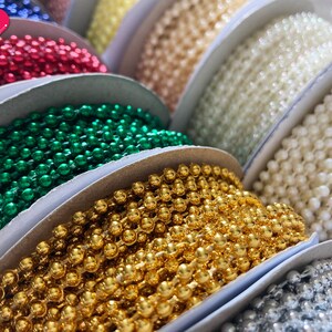 K&ouml;nnte beinhalten: Sortiment an Spulen mit bunten Perlen, darunter rot, gr&uuml;n, gold, wei&szlig; und silber. Die Perlen sind kreisf&ouml;rmig auf einer wei&szlig;en Oberfl&auml;che angeordnet.