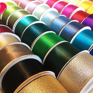 May include: A collection of colorful ribbon spools in various shades, including gold, black, brown, green, blue, pink, and purple. The ribbons have a satin finish, with some featuring a glittery texture. The spools are neatly arranged, showcasing the diverse color palette.
