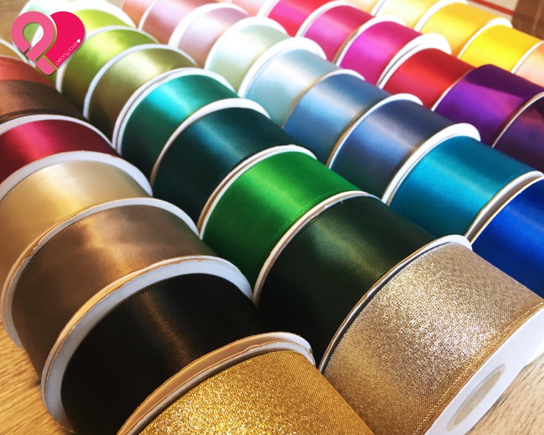May include: A collection of colorful ribbon spools in various shades, including gold, black, brown, green, blue, pink, and purple. The ribbons have a satin finish, with some featuring a glittery texture. The spools are neatly arranged, showcasing the diverse color palette.