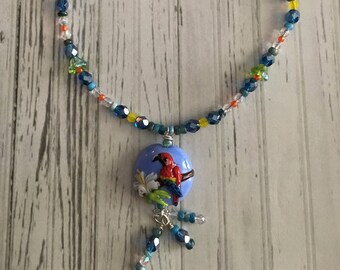 Puede incluir: Un collar de cadena plateada con un colgante de vidrio azul y blanco con un pájaro rojo. El colgante está rodeado de cuentas azules, verdes, amarillas y transparentes.