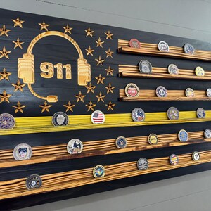May include: A wooden American flag with a yellow stripe and a 911 emblem with a headset. The flag has shelves for displaying challenge coins.
