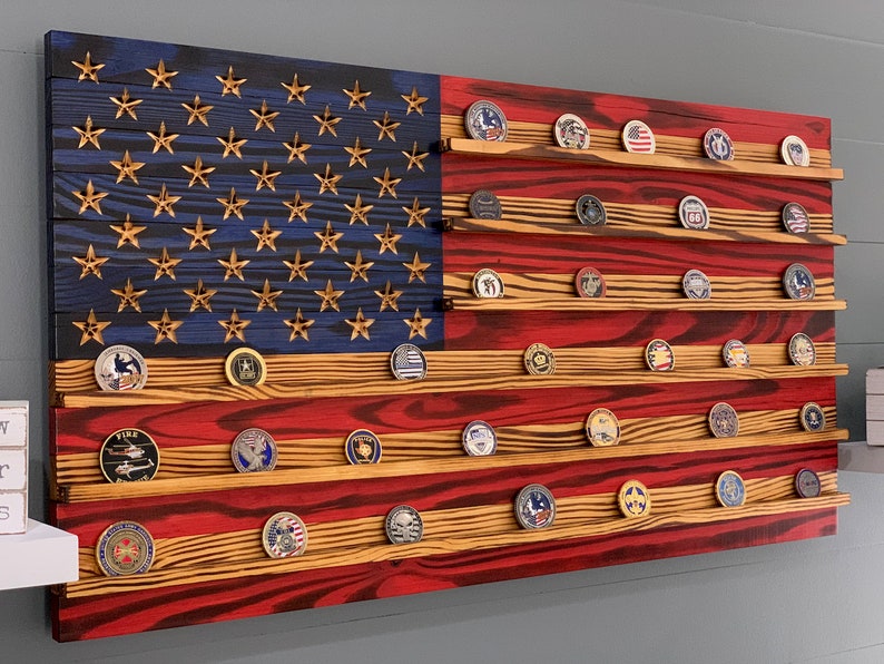 May include: A wooden American flag with a red, white, and blue design. The flag has shelves for displaying challenge coins. The shelves are filled with various challenge coins.
