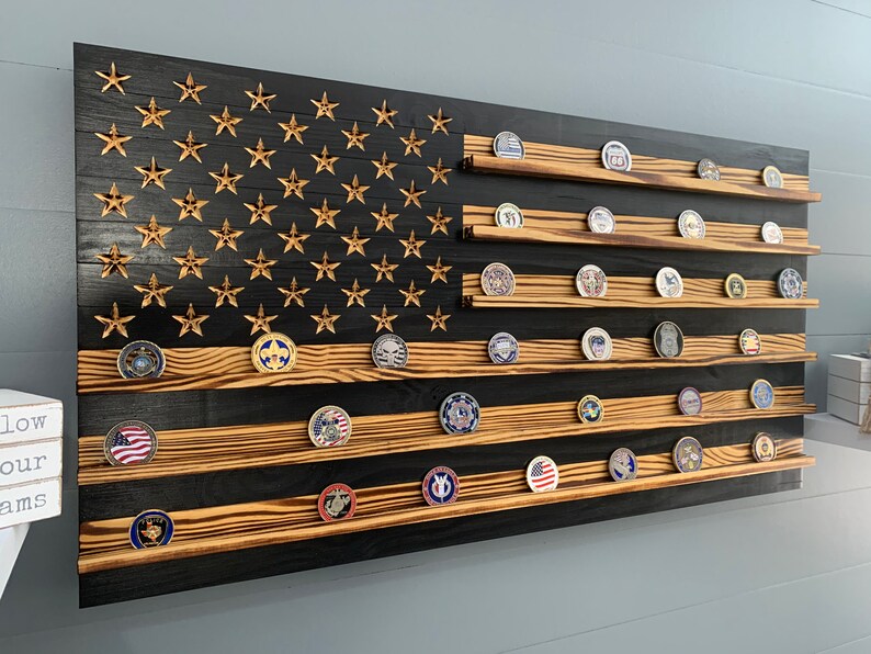 May include: A wooden American flag with a black and brown wood grain finish. The flag has 13 horizontal stripes and 50 stars. There are shelves on the flag to display challenge coins.