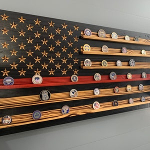May include: A wooden American flag with a red stripe, black background, and gold stars. The flag has multiple shelves for displaying challenge coins.