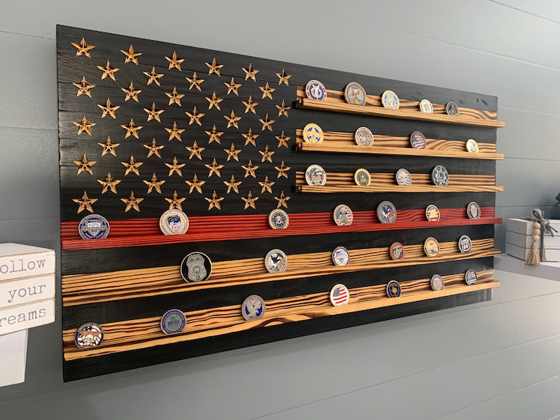 May include: A wooden American flag with a red stripe, black background, and gold stars. The flag has multiple shelves for displaying challenge coins.