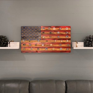 May include: A wooden American flag with red, white, and blue paint. The flag is decorated with small round metal discs, each with a different design. The flag is hanging on a wall above a white shelf with decorative items.