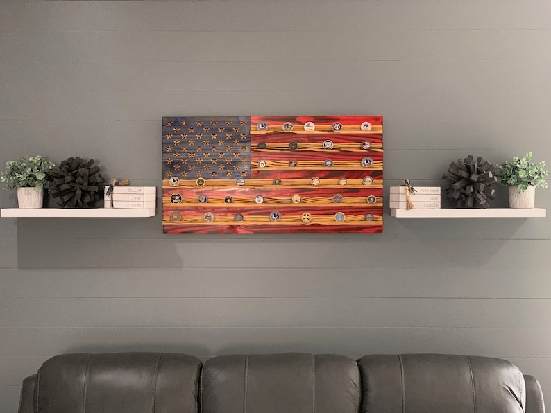 May include: A wooden American flag with red, white, and blue paint. The flag is decorated with small round metal discs, each with a different design. The flag is hanging on a wall above a white shelf with decorative items.