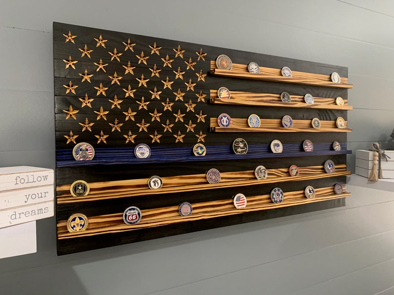 May include: A wooden American flag with a blue line, designed to hold challenge coins. The flag is stained black with a burnt wood finish and has gold stars. The flag has multiple shelves to display the coins.
