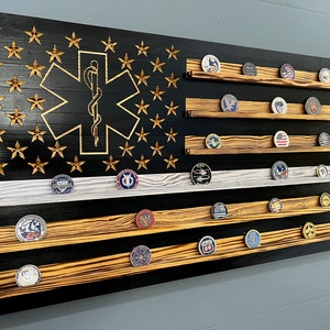May include: A black and white wooden American flag with a gold star of life and a medical caduceus symbol. The flag has five shelves for displaying challenge coins.