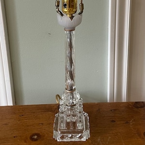 May include: A clear glass table lamp with a crystal base and a gold-toned metal shade. The lamp has a clear glass bulb with a filament.