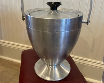May include: A silver ice bucket with a lid and a handle. The bucket has a round base and a flared top. The lid has a black knob.