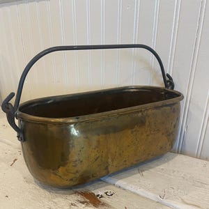May include: An antique brass bucket with a black metal handle. The bucket has a rectangular shape with rounded edges and a slightly tarnished finish, suggesting age and use. The handle is attached to the sides with decorative metal loops. The bucket sits on a distressed white wooden surface.