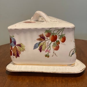 May include: White ceramic cheese dish with a lid and a handle. The dish is decorated with a floral pattern of red strawberries and green leaves.