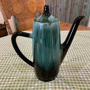 Könnte beinhalten: Eine schwarze und grüne Kaffeekanne aus Keramik mit Deckel. Die Kanne hat einen Ausgießer und einen Griff. Die Kanne steht auf einer grün-weiß karierten Tischdecke.