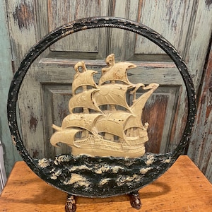 May include: A cast iron fireplace screen featuring a ship sailing on the waves. The screen is painted in black and cream colors and has a decorative border around the edge. It is on a wooden surface.