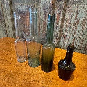 May include: Four antique glass bottles with different shapes and colours. The bottles are arranged in a row on a wooden surface.
