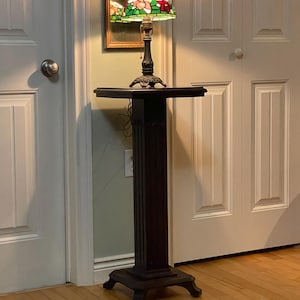 May include: A tall, dark brown wooden pedestal stand with a square top and base, featuring four feet. A stained glass table lamp with a floral pattern in red, white, and green sits on the pedestal, emitting a warm light from its metal base.