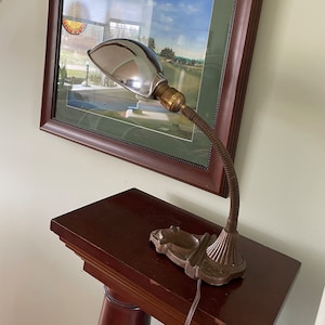 Peut inclure: Une lampe de bureau vintage en chrome et en laiton avec un bras flexible en col de cygne et une base décorative. La lampe est posée sur une table en bois devant un tableau encadré.