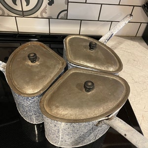 May include: Three vintage enamelware pots with blue and white speckled paint and silver lids. The pots have black handles and knobs.