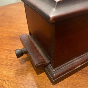 May include: A dark stained wooden box with a pull-out drawer and a small, round knob. The box has a rectangular shape with a decorative top edge. The wood grain is visible, and the finish is slightly worn.