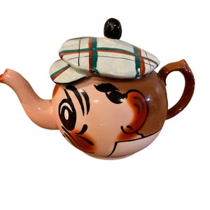 May include: A ceramic teapot shaped like a cartoon face with a plaid hat. The teapot is pink with brown accents and black eyes. The teapot has a brown handle and a spout.