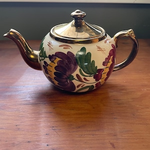 Puede incluir: Una tetera blanca con tapa dorada y diseño floral en tonos de morado, verde y amarillo. La tetera está sobre una superficie de madera.