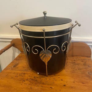 May include: A black and cream-colored ice bucket with a lid and metal handle. The bucket has a decorative metal frame with leaf and swirl designs. The lid has a small, decorative knob. The ice bucket is sitting on a wooden surface.