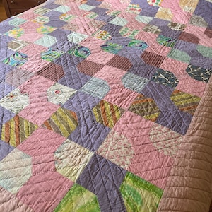 May include: A patchwork quilt in pink and purple hues, featuring a geometric design of hexagons and diamonds. The quilt showcases a variety of fabric patterns and textures. It is likely made of cotton and has a quilted finish, suitable for a double bed.