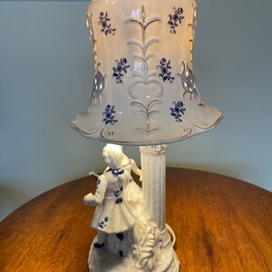 May include: A white porcelain table lamp with a blue and white patterned shade. The lamp base features a couple in a romantic pose.