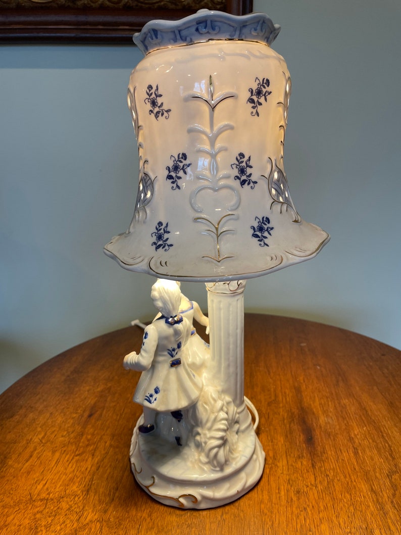 May include: A white porcelain table lamp with a blue and white patterned shade. The lamp base features a couple in a romantic pose.