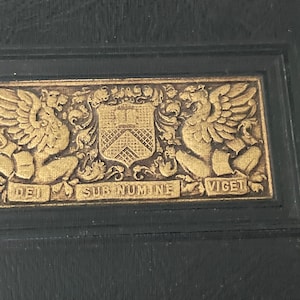 May include: A gold embossed crest with two winged creatures on either side of a shield. The crest is surrounded by a black frame and the text "DEI SUB NUMINE VIGET" is written below the crest.