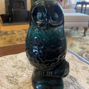 May include: A ceramic owl figurine in shades of dark teal and black. The owl has large, round eyes and is perched on a small base. The glossy finish reflects light, highlighting the textured surface. A decorative item for home decor.