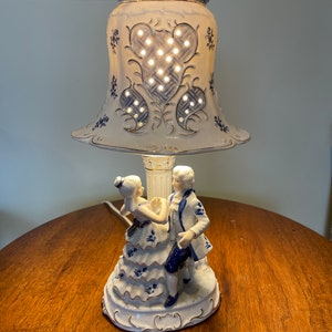 May include: A white porcelain table lamp with a blue and gold design. The lamp features a couple dancing on the base and a bell-shaped shade with a cut-out design.