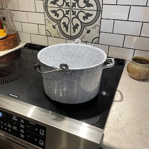 May include: A large, white enamel pot with a speckled gray finish and a black handle sits on a black electric stovetop. The stovetop has a digital display with the word "INDUCTION" printed on it.