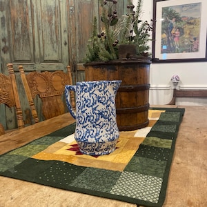 Peut inclure: Un pichet en céramique bleu et blanc avec un motif floral posé sur un chemin de table en patchwork vert et jaune. Le pichet est sur une table en bois avec une chaise en bois en arrière-plan.
