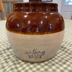 Puede incluir: Una olla de barro esmaltada en marrón y blanco con tapa. La olla tiene un diseño de corona con el número "2" dentro de la corona.