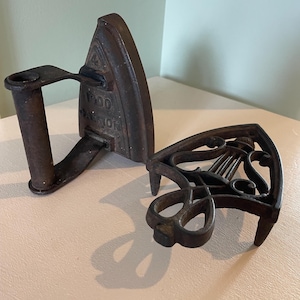 May include: A dark brown cast iron sad iron with a handle and the text 1900 OAKTON embossed on the side, accompanied by a matching dark brown cast iron trivet featuring an openwork design with a central lyre shape and three pointed feet.