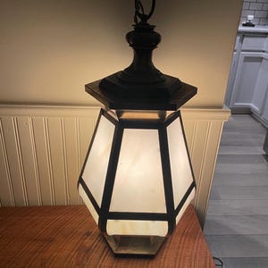 May include: A vintage, octagonal, frosted glass pendant light fixture with a black metal frame and a decorative bronze top. The light is hanging from a chain and is on a wooden table.