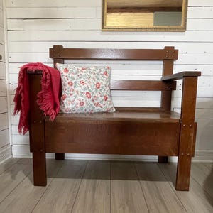 May include: A brown wooden bench with a high backrest and armrests. A square throw pillow with a white background and a red and green floral pattern sits on the seat. A red fringed throw blanket is draped over the left armrest of the bench.