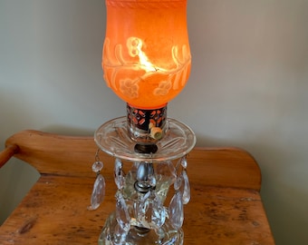 Könnte beinhalten: Eine Vintage-Tischlampe mit einem Sockel aus klarem, facettiertem Glas und einem orangefarbenen Glasschirm. Der Schirm hat Blumen- und Blättermuster und einen gewellten Rand. Klare Kristallprismen hängen von der Mitte der Lampe.